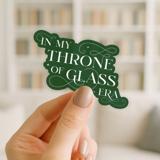 Hand holding a green decorative sign with 'In my Throne of Glass Era' text against a blurred bookshelf background.
