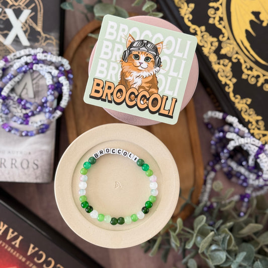 Beaded bracelet with 'Broccoli' text on a wooden coaster, surrounded by decorative items.
