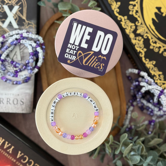 Beaded bracelet in a box with 'We Do Not Eat Our Allies' tag, surrounded by books and plants.