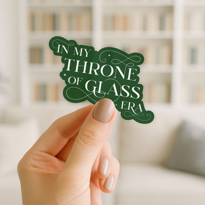 Hand holding a green decorative sign with 'In my throne of glass era' text against a blurred bookshelf background.