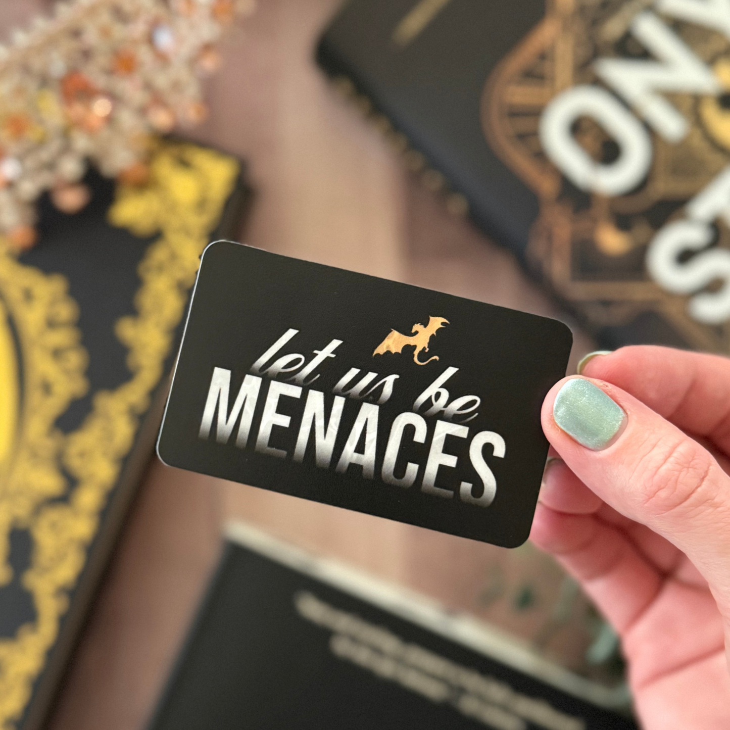 Hand holding a black card with 'let us be MENACES' text, with decorative items in the background.