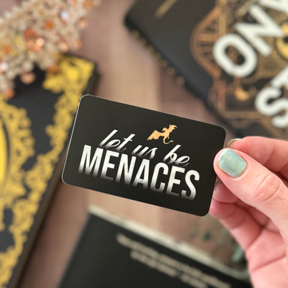 Hand holding a black card with 'let us be MENACES' text, with decorative items in the background.