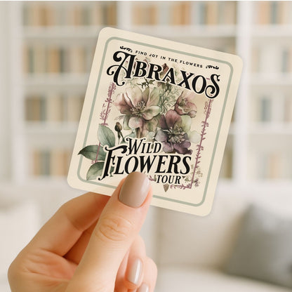 Hand holding a card with 'Abraxos Wild Flowers Tour' design against a blurred background