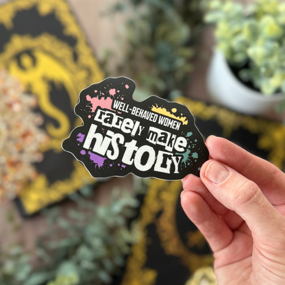 Hand holding a sticker with text 'Well-behaved women rarely make history' against a blurred background.