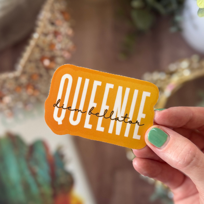 Hand holding a yellow sticker with 'QUEENIE diem bellator' text against a blurred background