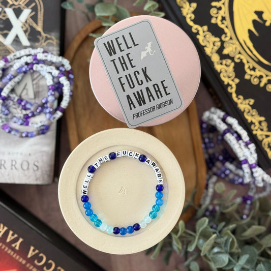 Bracelet with 'Well The F**k Aware' text in a pink box with a card, surrounded by books and plants.