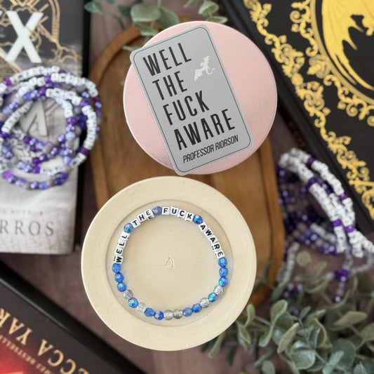 Bracelet with 'Well The F**k Aware' text on a pink and beige display stand, surrounded by books and plants.