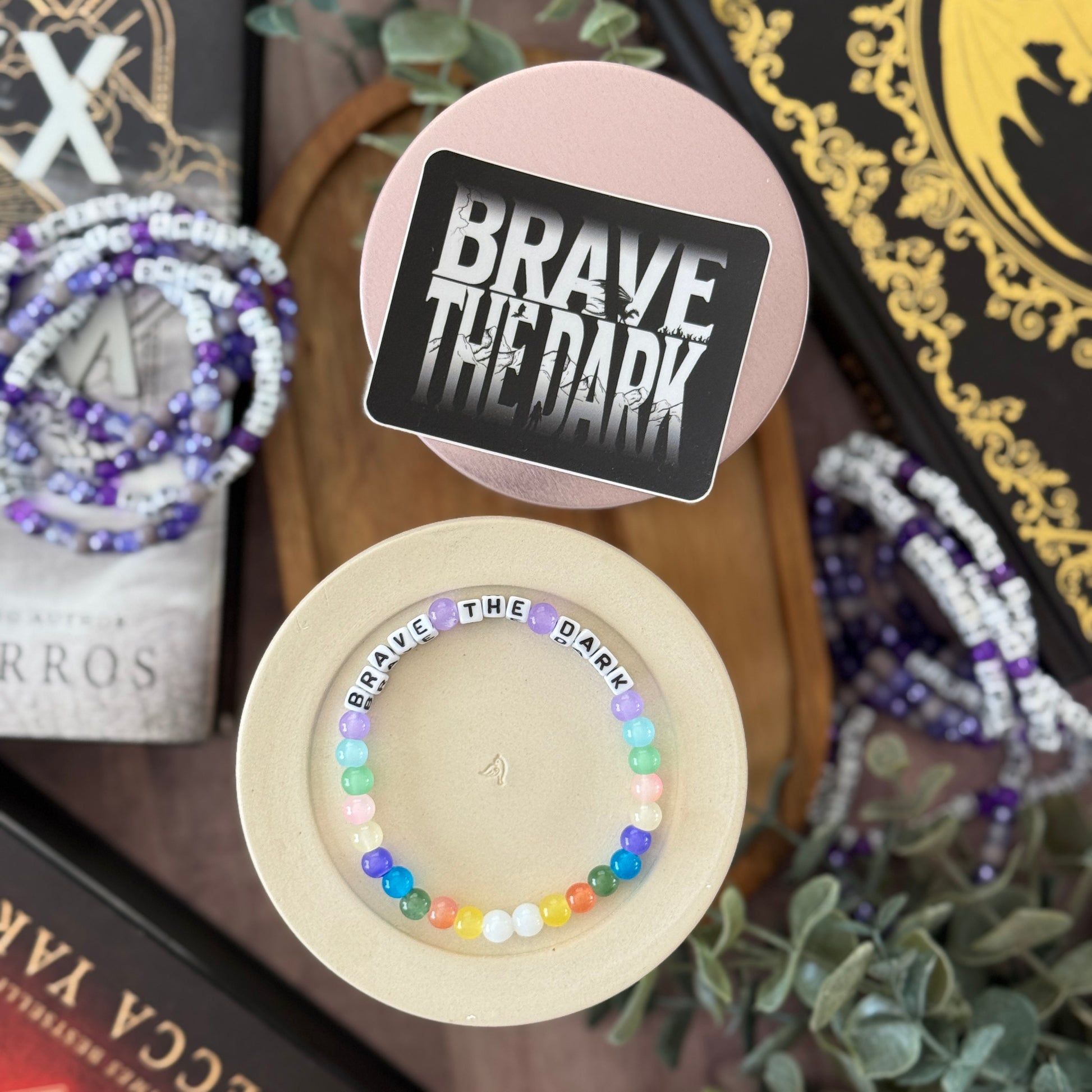 Colorful beaded bracelet with 'BRAVE THE DARK' text on a wooden stand, surrounded by books and plants.