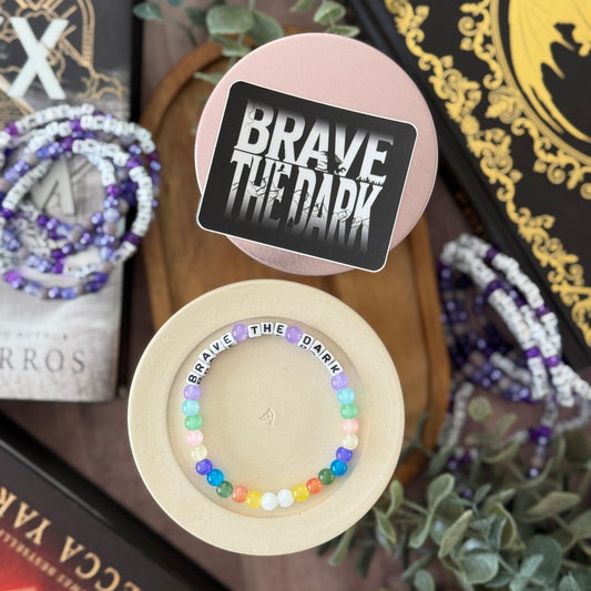 Colorful beaded bracelet with 'BRAVE THE DARK' text on a wooden stand, surrounded by books and plants.