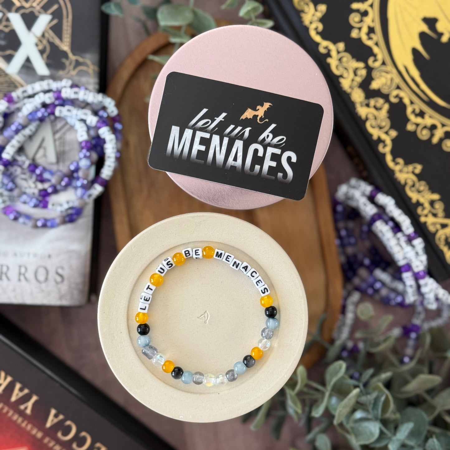 Beaded bracelet with 'let us be MENACES' message in a display box, surrounded by books and plants.