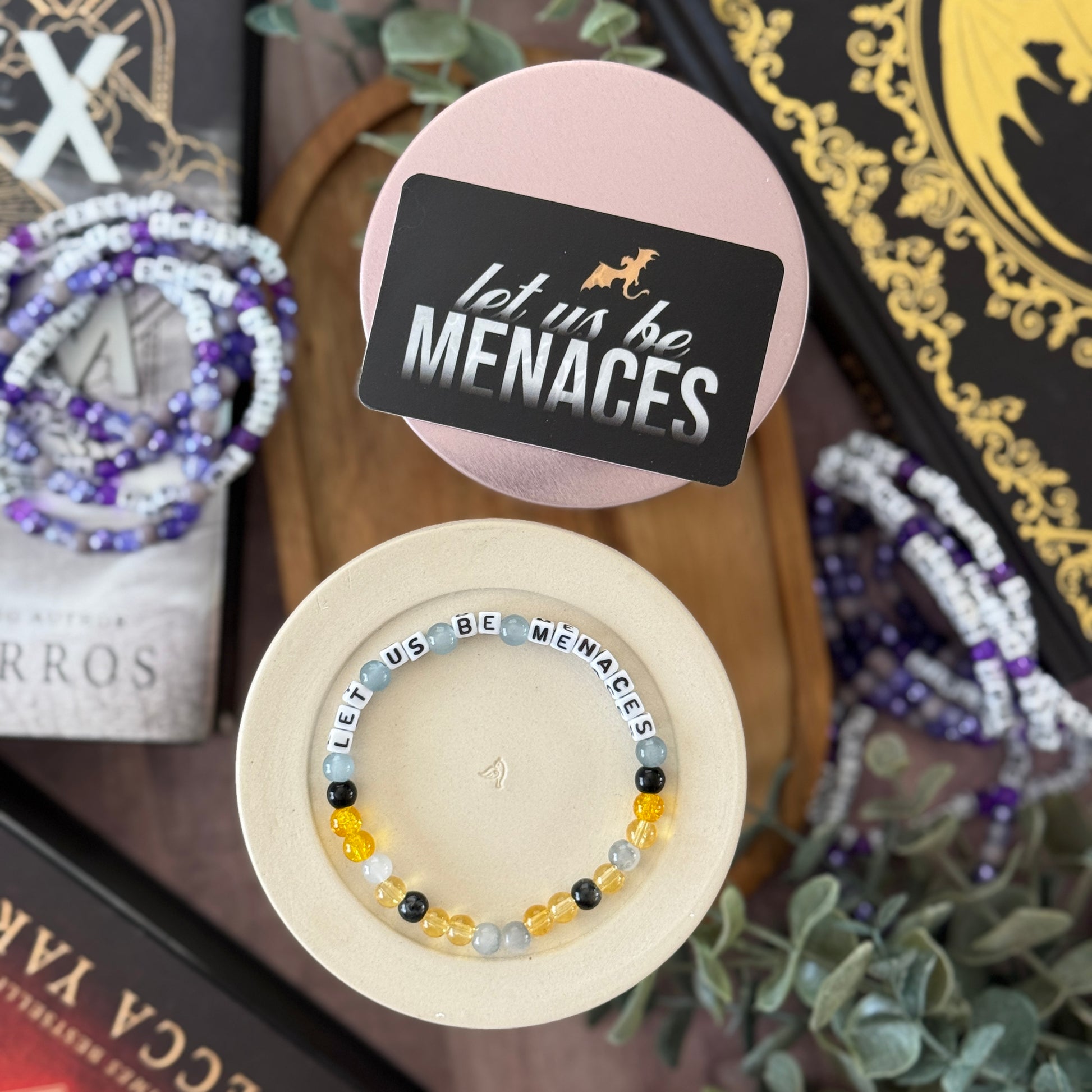 Bracelet with 'let us be MENACES' text on a wooden stand, surrounded by books and plants.
