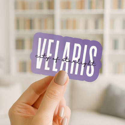 Hand holding a purple sticker with 'VELARIS city of starlight' text against a blurred indoor background.