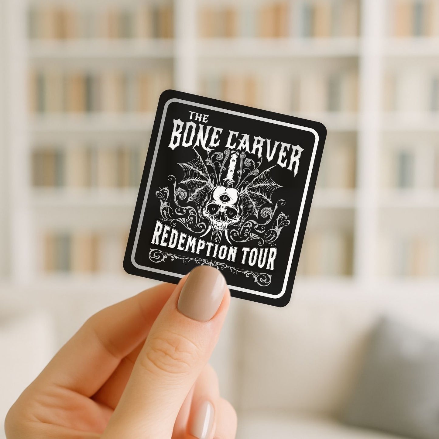 Hand holding a 'The Bone Carver Redemption Tour' sticker with a blurred bookshelf background