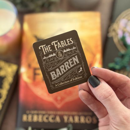 Hand holding a card titled 'The Fables of the Barren' with a book in the background