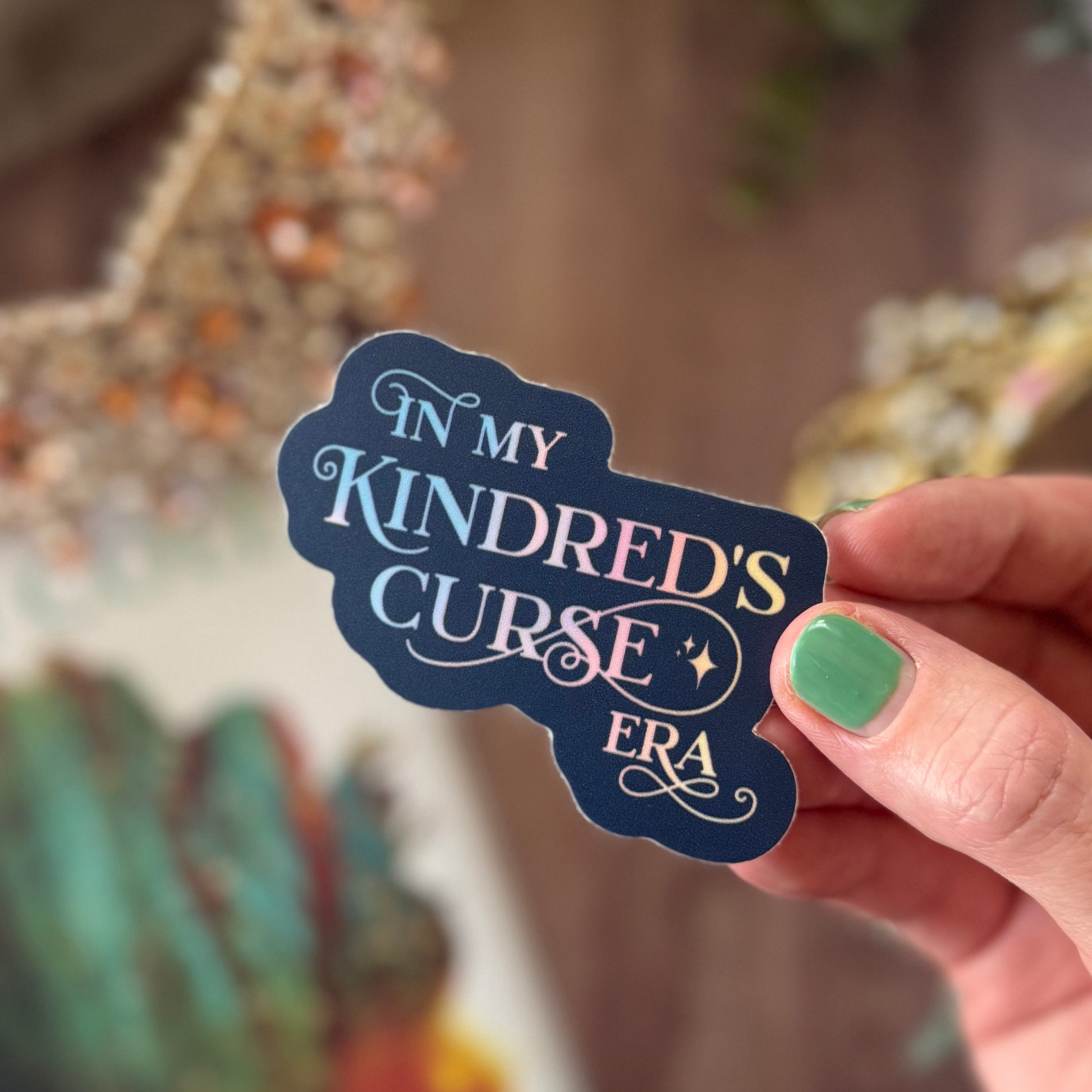 Hand holding a sticker with 'In My Kindred's Curse Era' text against a blurred background