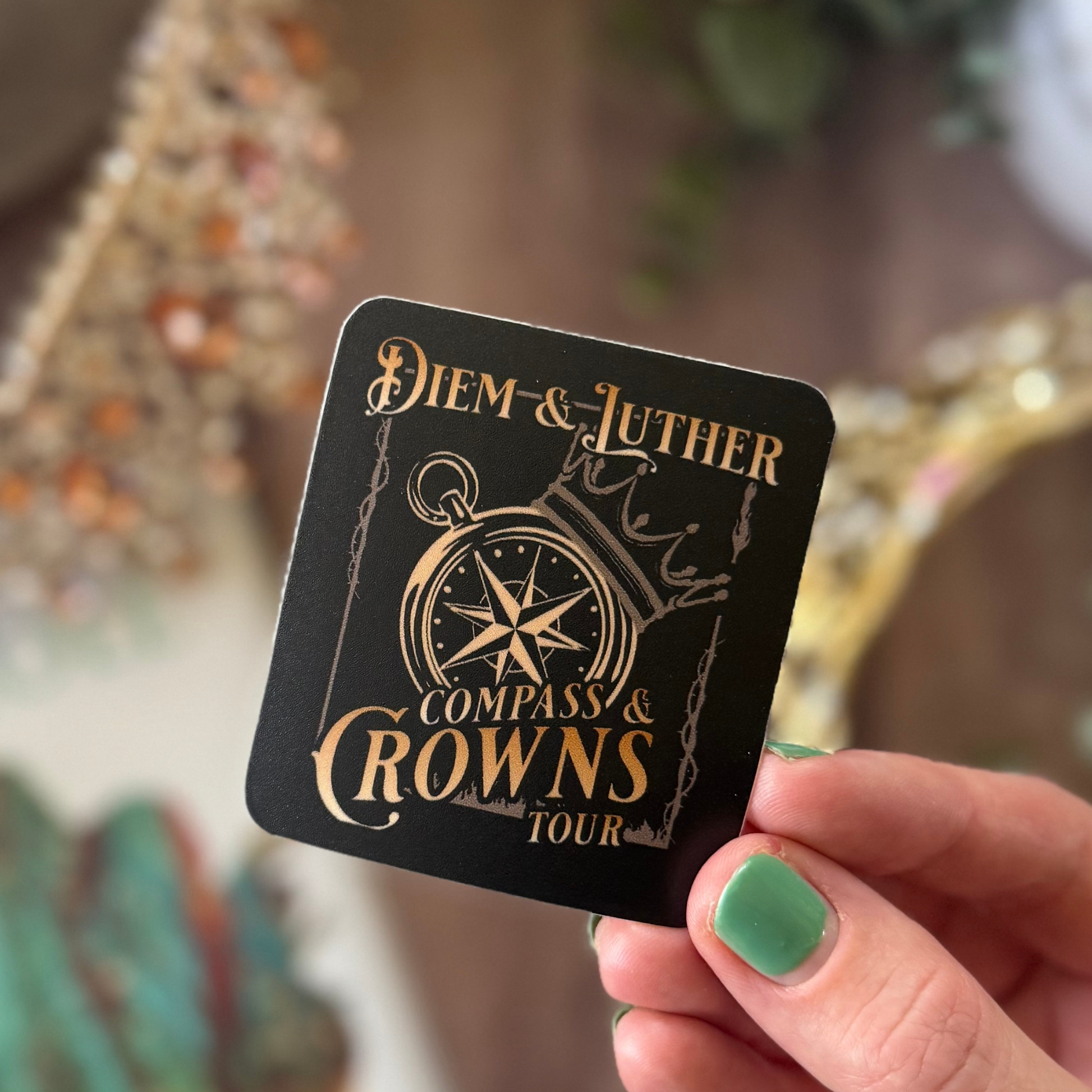 Hand holding a black card with gold text and design, featuring 'Diem & Luther Compass & Crowns Tour'.