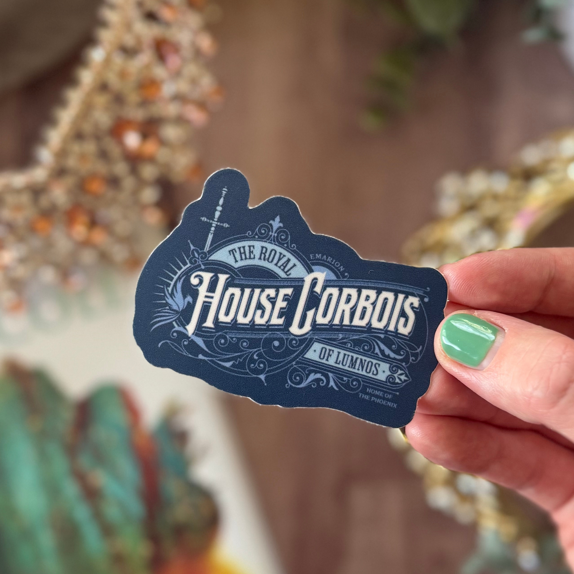 Hand holding a 'The Royal House Corbois' sticker with decorative text.
