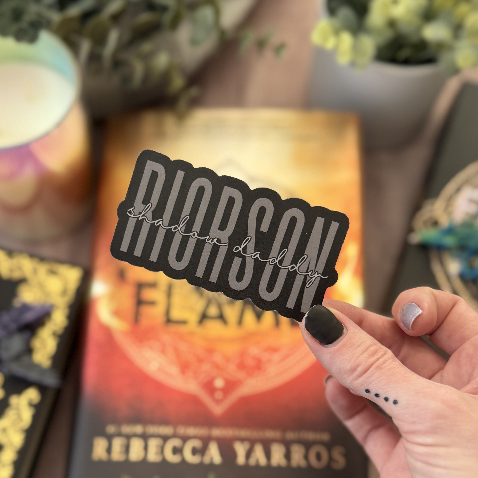 Hand holding a 'Diorison' sticker over a book titled 'Flame' by Rebecca Yarros with a blurred background.