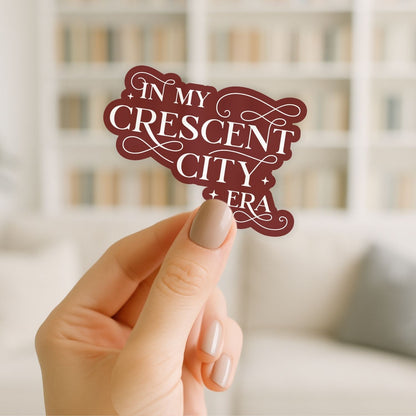 Hand holding a decorative sign with 'In My Crescent City Era' text against a blurred indoor background.