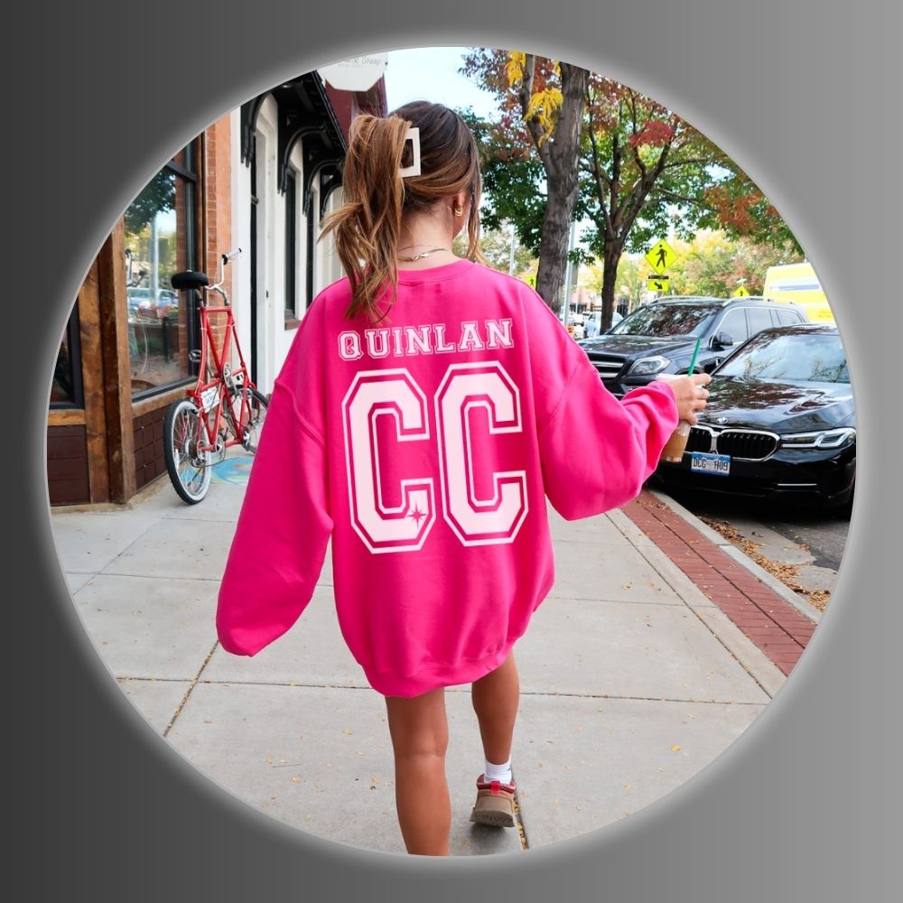Girl wearing a pink sweatshirt with 'QUINLAN CC' on a city street.
