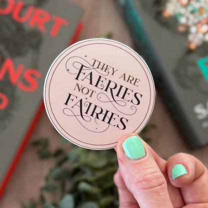 Hand holding a round sticker with text 'THEY ARE FAERIES NOT FAERIES' against a blurred background.