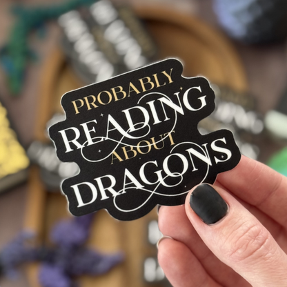 Hand holding a sticker with 'Probably Reading About Dragons' text against a blurred bookshelf background.