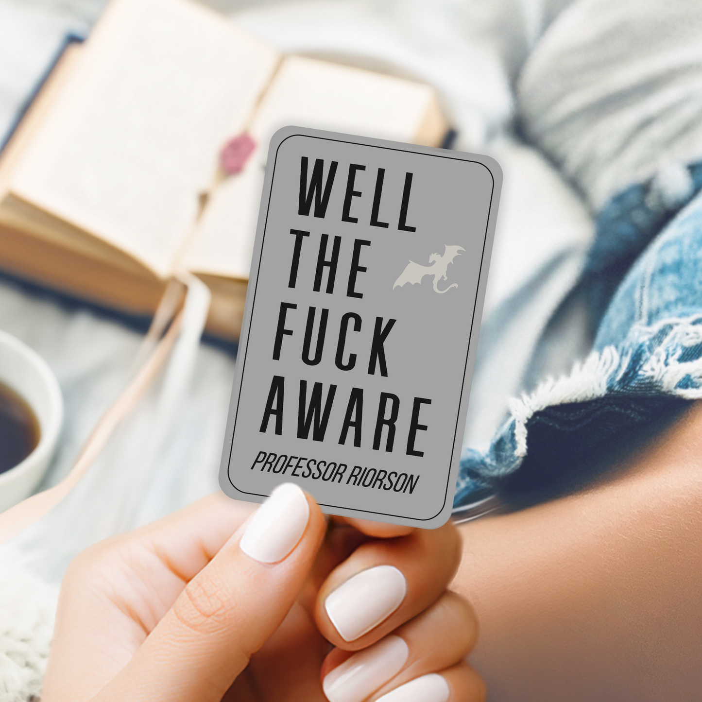 Person holding a card with text 'Well the fuck aware' by Professor Riorson, with a blurred background of books and a cup.