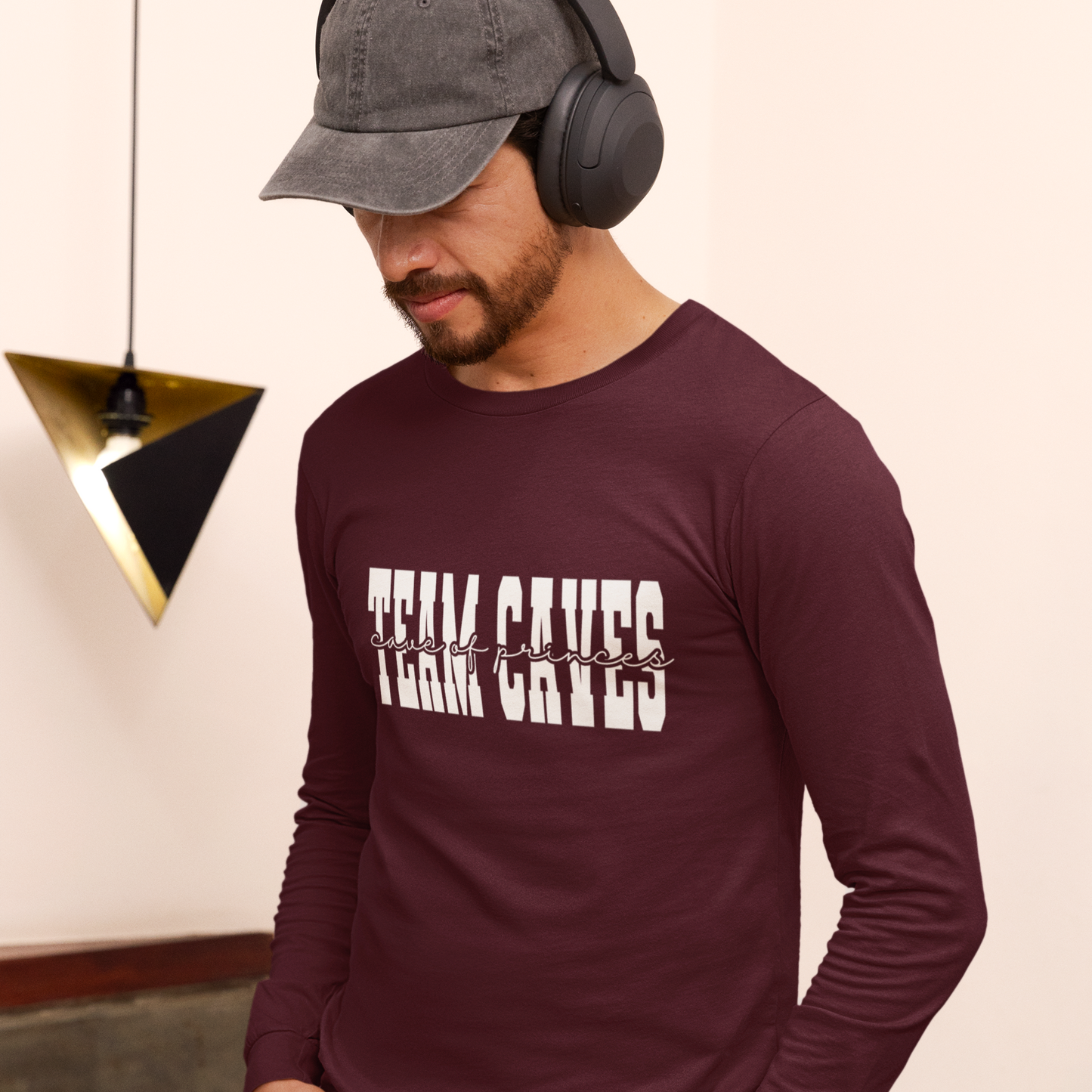 Man wearing a maroon long-sleeve shirt with 'Team Caves' text, gray cap, and headphones indoors.