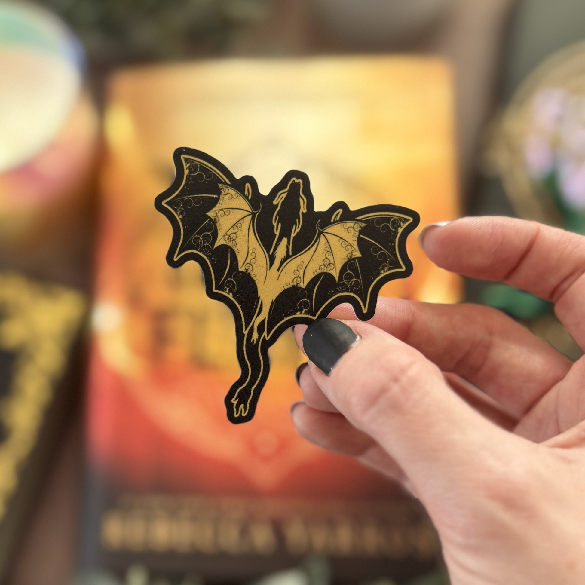 Hand holding a decorative bat sticker with a blurred bookshelf in the background