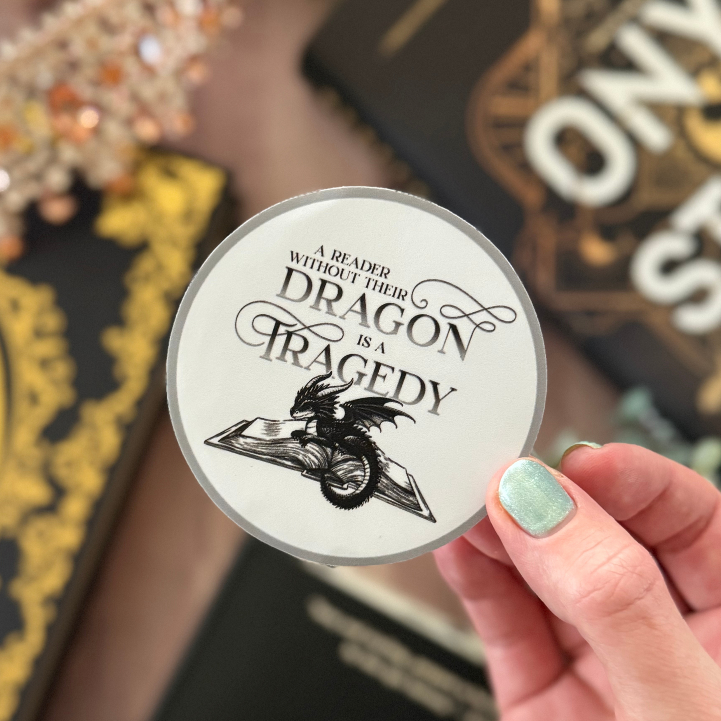 Circular sticker with dragon design and text held by a hand, with books in the background